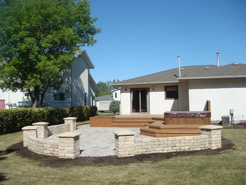 Treated brown deck with built-in hot tub and planters. Wide staircase leading onto Roman paver patio with Quarry Stone pillars and seating walls.