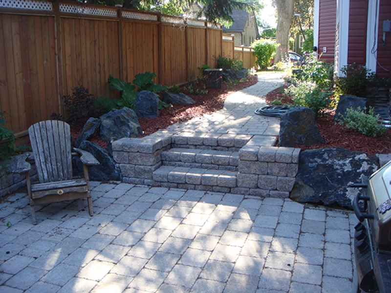 Complete yard renovation. Roman paving stones and retaining walls. Plantings and boulders surrounded by red wood mulch. Treated brown fence with white lattice top (Retaining Walls)