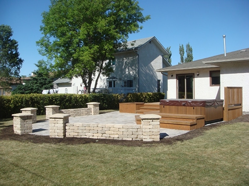 Treated brown deck with built-in hot tub and planters. Wide staircase leading onto Roman paver patio with Quarry Stone pillars and seating walls.